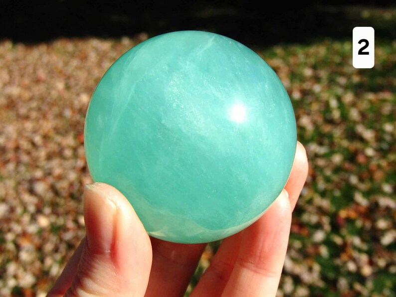 May include: A smooth, round, light teal-colored sphere. The sphere is held in a hand, with the background out of focus. The sphere has a glossy finish and appears to be made of a translucent material. The number "2" is in the upper right corner.