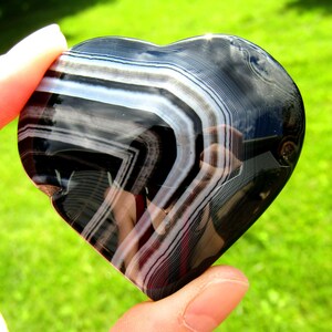 May include: A polished heart-shaped stone featuring distinct, alternating bands of deep black and translucent white. The surface is smooth and reflective, showcasing natural mineral layers that create a sharp, geometric pattern across the curved form.