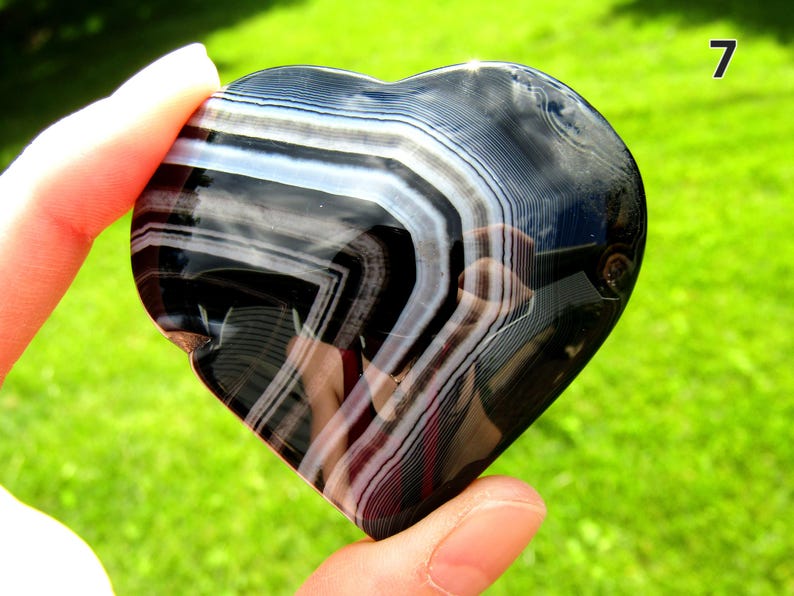 May include: A polished heart-shaped stone featuring distinct, alternating bands of deep black and translucent white. The surface is smooth and reflective, showcasing natural mineral layers that create a sharp, geometric pattern across the curved form.