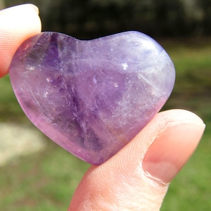 Puede incluir: Un cristal de amatista en forma de coraz&oacute;n, sostenido entre dos dedos. La piedra pulida muestra tonos de p&uacute;rpura y lavanda, con variaciones naturales en color y textura. El cristal es liso y redondeado, reflejando la luz.