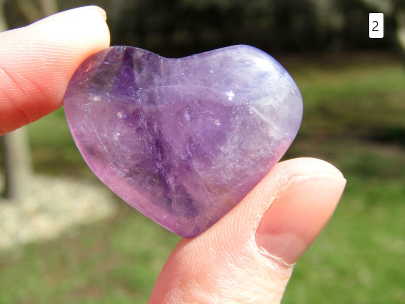 Puede incluir: Un cristal de amatista en forma de coraz&oacute;n, sostenido entre dos dedos. La piedra pulida muestra tonos de p&uacute;rpura y lavanda, con variaciones naturales en color y textura. El cristal es liso y redondeado, reflejando la luz.
