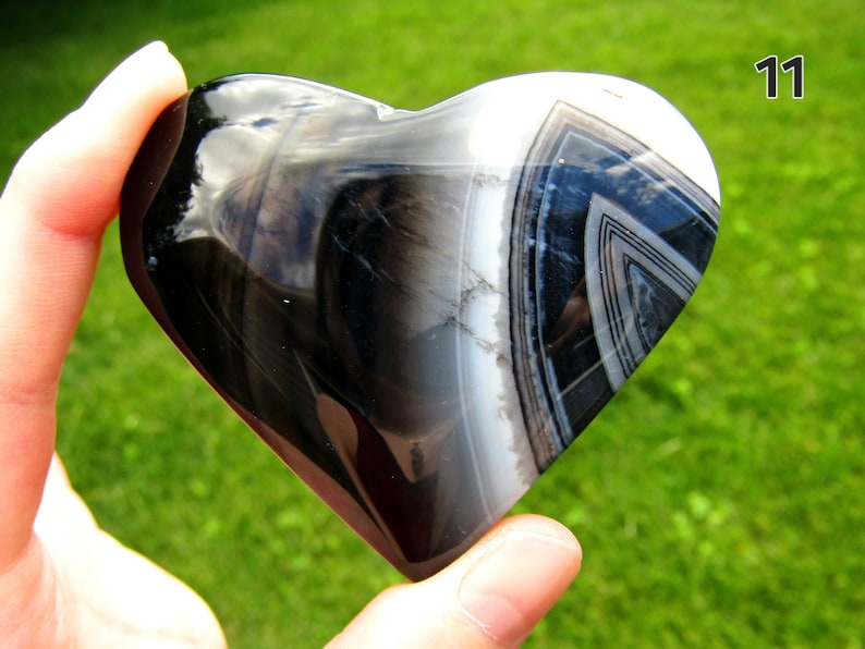 May include: A polished heart-shaped stone featuring a smooth, glossy surface with distinct bands of black, white, and gray. The stone displays a natural layered pattern with a prominent triangular design on the right side and the number 11.