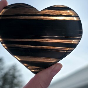 May include: A heart-shaped stone with a polished surface. The stone features alternating bands of black and gold, creating a striped pattern. The heart is held up against a bright, sunny background.