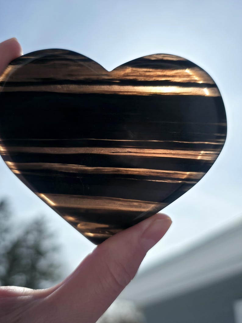 May include: A heart-shaped stone with a polished surface. The stone features alternating bands of black and gold, creating a striped pattern. The heart is held up against a bright, sunny background.