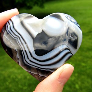 May include: A polished heart-shaped stone featuring distinct bands of black, white, and translucent gray. The surface is smooth and reflective, showcasing natural concentric layers and patterns throughout the stone. The number 1 is visible.