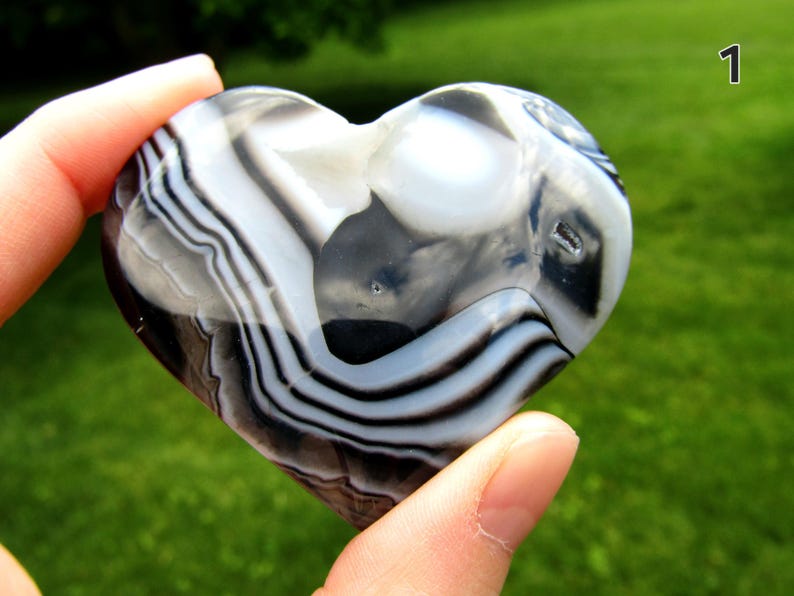May include: A polished heart-shaped stone featuring distinct bands of black, white, and translucent gray. The surface is smooth and reflective, showcasing natural concentric layers and patterns throughout the stone. The number 1 is visible.