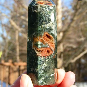 May include: A green and orange gemstone obelisk. The polished stone features a mix of dark green and white patterns with orange-brown inclusions. The obelisk is held in a hand, with a natural outdoor background.