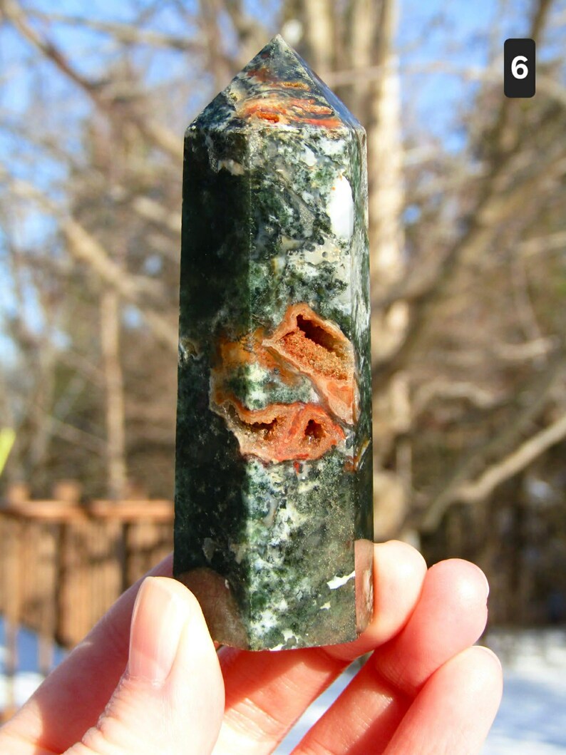 May include: A green and orange gemstone obelisk. The polished stone features a mix of dark green and white patterns with orange-brown inclusions. The obelisk is held in a hand, with a natural outdoor background.