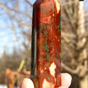 May include: A polished gemstone obelisk with a pointed top. The stone features a mix of red, brown, white, and green colors. The obelisk is held in a hand against a blurred outdoor background.