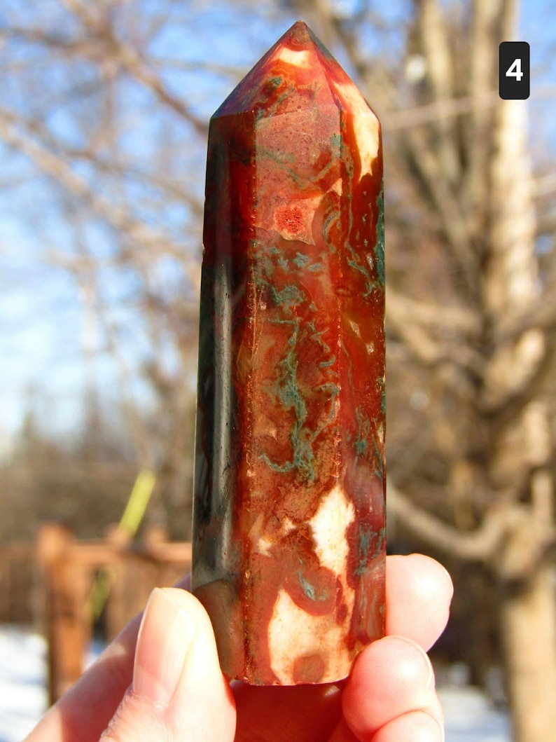 May include: A polished gemstone obelisk with a pointed top. The stone features a mix of red, brown, white, and green colors. The obelisk is held in a hand against a blurred outdoor background.