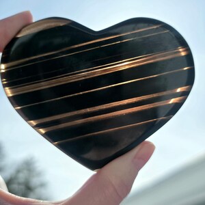 May include: A heart-shaped, polished stone with a dark, almost black base color. The stone is accented with parallel, golden-brown stripes. The heart is held up against a bright, sunny sky.