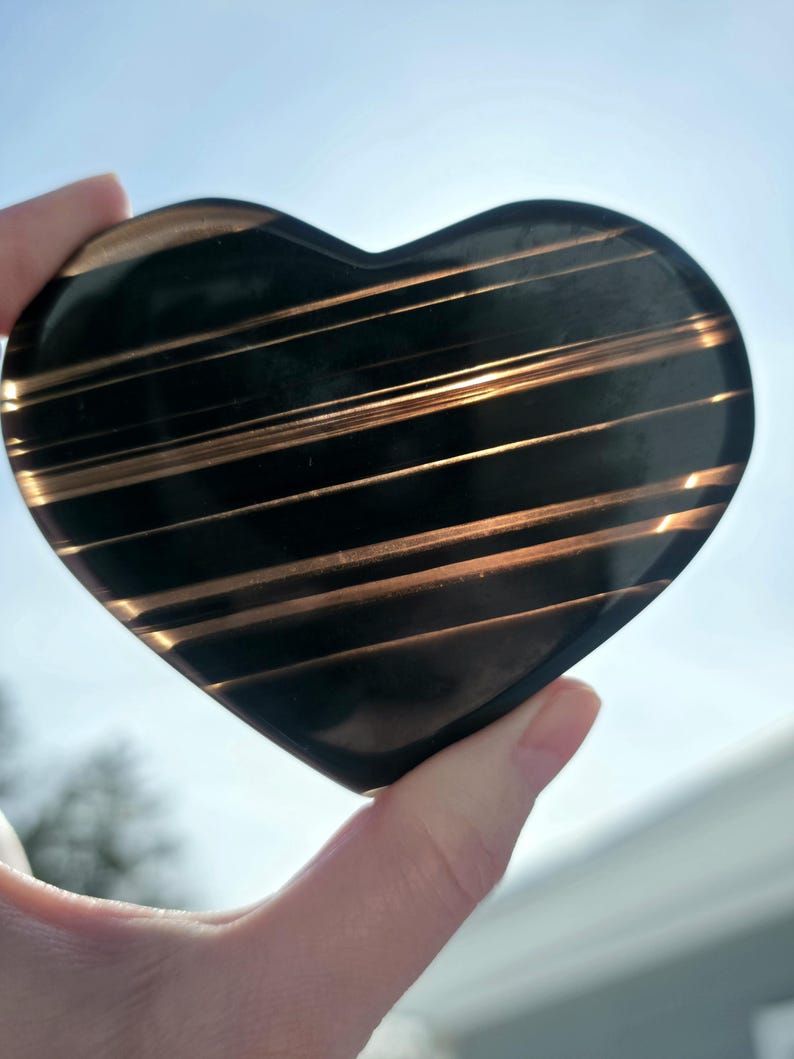 May include: A heart-shaped, polished stone with a dark, almost black base color. The stone is accented with parallel, golden-brown stripes. The heart is held up against a bright, sunny sky.