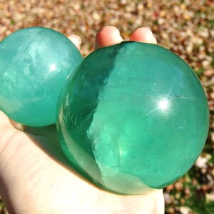 May include: Two translucent, teal-colored spheres are held in a hand. The spheres have a smooth, polished surface and appear to be made of a crystalline material. The background is blurred, with hints of autumn leaves.
