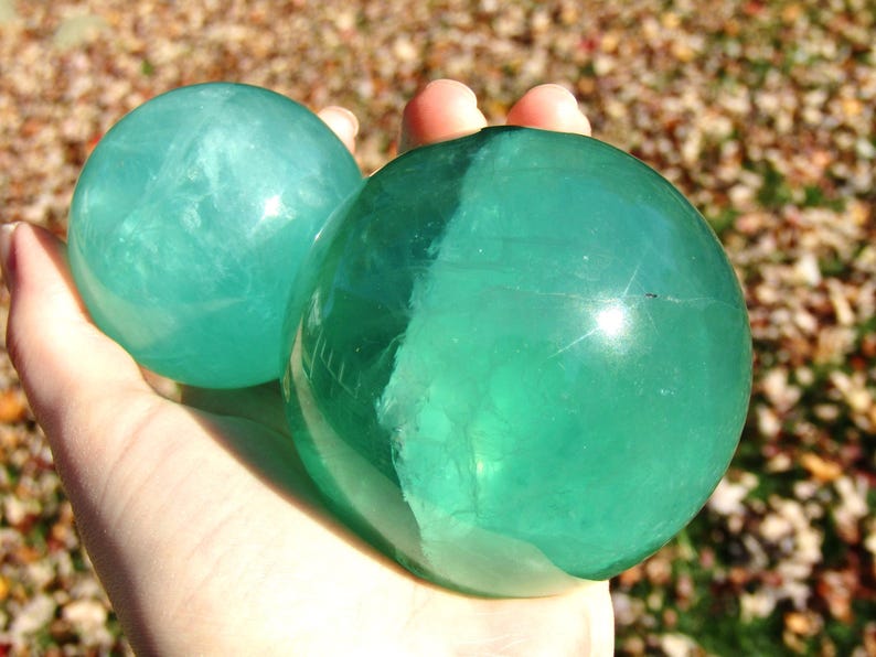 May include: Two translucent, teal-colored spheres are held in a hand. The spheres have a smooth, polished surface and appear to be made of a crystalline material. The background is blurred, with hints of autumn leaves.