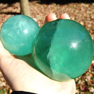 May include: Two polished, translucent spheres in shades of teal and green are held in a hand. The spheres have a smooth, reflective surface, with internal veining visible. The background is blurred, suggesting an outdoor setting.