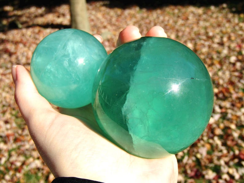 May include: Two polished, translucent spheres in shades of teal and green are held in a hand. The spheres have a smooth, reflective surface, with internal veining visible. The background is blurred, suggesting an outdoor setting.