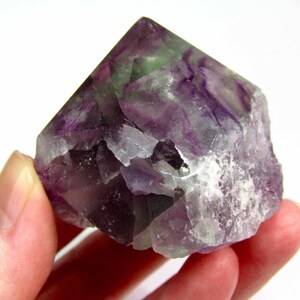 May include: A purple, green, and white fluorite crystal point. The crystal is a natural, raw, and unpolished specimen. It has a pointed top and a rough, irregular base.