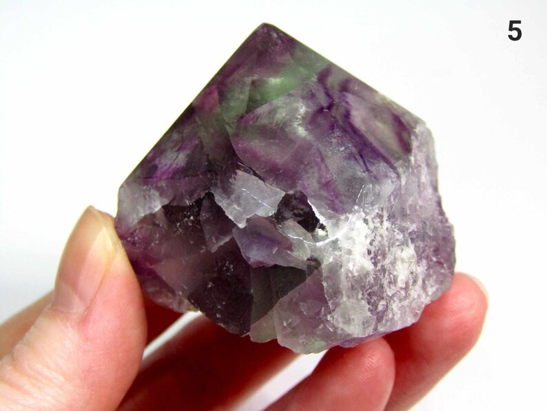 May include: A purple, green, and white fluorite crystal point. The crystal is a natural, raw, and unpolished specimen. It has a pointed top and a rough, irregular base.