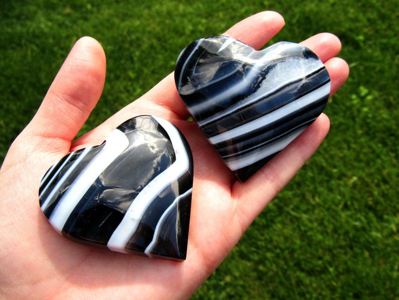May include: Two polished heart-shaped stones featuring a smooth, glossy finish with distinct, alternating bands of opaque black and bright white. The stones exhibit a natural, layered appearance with curved, flowing lines that follow the heart shape.