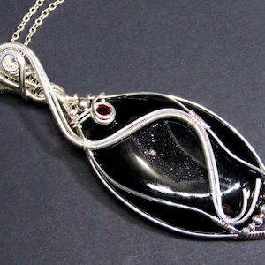 Op de afbeelding: Een zilveren draadgewikkelde hanger met een zwarte edelsteen met een rode edelsteen accent. De hanger is bevestigd aan een zilveren ketting.