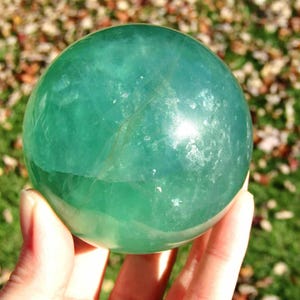 May include: A translucent, green fluorite sphere. The polished gemstone is held in a hand, with a blurred background of green grass and fallen leaves. The sphere has internal veining and a smooth, reflective surface. A white number 5 is in the upper right corner.
