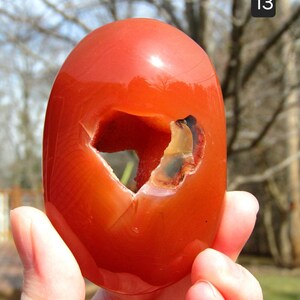 K&ouml;nnte beinhalten: Ein polierter, oval geformter, orangeroter Stein mit einer gro&szlig;en, unregelm&auml;&szlig;igen H&ouml;hlung. Der Stein wird in einer Hand gehalten, mit der Nummer "13" oben rechts. Die Oberfl&auml;che des Steins ist glatt und reflektierend.