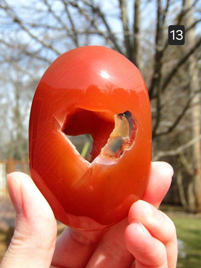 K&ouml;nnte beinhalten: Ein polierter, oval geformter, orangeroter Stein mit einer gro&szlig;en, unregelm&auml;&szlig;igen H&ouml;hlung. Der Stein wird in einer Hand gehalten, mit der Nummer "13" oben rechts. Die Oberfl&auml;che des Steins ist glatt und reflektierend.