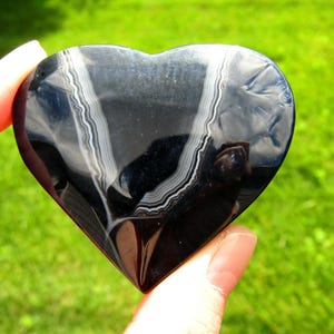 May include: A polished heart-shaped stone with a smooth, glossy black surface featuring two distinct, curved white banded patterns running vertically across the center. The number 2 is visible in the upper right corner of the image.