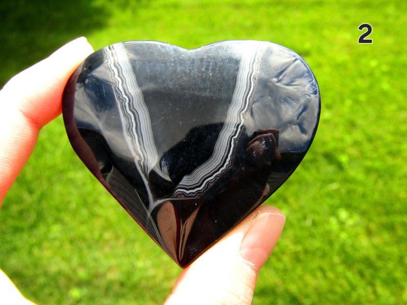 May include: A polished heart-shaped stone with a smooth, glossy black surface featuring two distinct, curved white banded patterns running vertically across the center. The number 2 is visible in the upper right corner of the image.