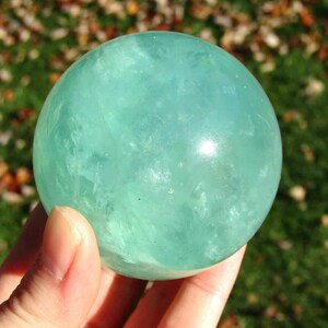 May include: A translucent, light teal fluorite sphere. The crystal ball is held in a hand, with a blurred background of green grass and autumn leaves. The sphere is a decorative item, possibly for display or meditation.
