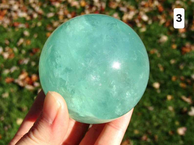 May include: A translucent, light teal fluorite sphere. The crystal ball is held in a hand, with a blurred background of green grass and autumn leaves. The sphere is a decorative item, possibly for display or meditation.
