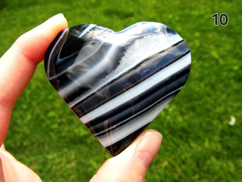 May include: A heart-shaped polished stone featuring alternating bands of black and white with a small crystalline inclusion near the top. The number 10 is printed in the upper right corner of the image against a natural green background.