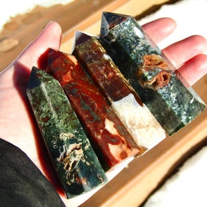 May include: Four polished gemstone obelisks in shades of green, red, brown, and white are held in a hand. The obelisks have a rectangular shape with a pointed top. The gemstones have unique patterns and textures.