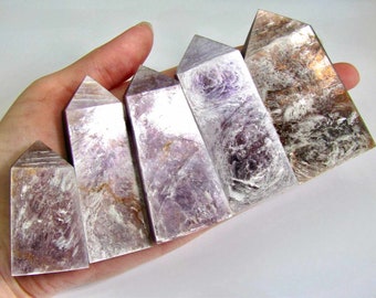 May include: Six iridescent purple and white lepidolite crystal points of varying sizes, arranged in a row on a hand.