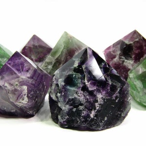 May include: A group of seven fluorite crystals in various shades of green, purple, and black. The crystals are all pointed and have a natural, rough texture.