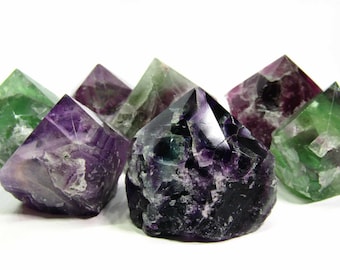 May include: A group of seven fluorite crystals in various shades of green, purple, and black. The crystals are all pointed and have a natural, rough texture.