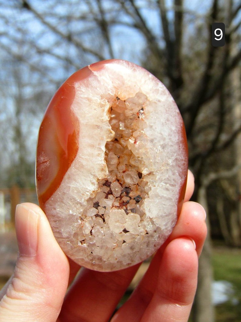 K&ouml;nnte beinhalten: Ovale Achat-Geode mit polierter, rotbrauner Au&szlig;enseite und einer mit funkelnden Quarzkristallen gef&uuml;llten H&ouml;hlung. Die Geode wird in einer Hand gehalten und zeigt ihre nat&uuml;rliche Sch&ouml;nheit und einzigartige Kristallstruktur.