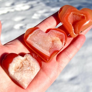 May include: Three heart-shaped agate stones in shades of orange and white are held in a hand. The stones have a polished, smooth exterior and a crystalline interior. The background is a blurred, snowy landscape.