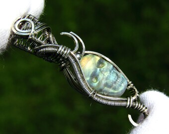 May include: A handcrafted pendant featuring a skull-shaped gemstone encased in intricate silver wire. The gemstone displays iridescent blue and green hues. The pendant is held by a gloved hand against a blurred green background.