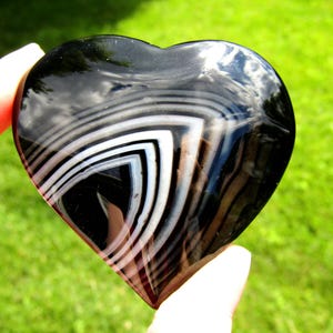 May include: A polished heart-shaped stone featuring a smooth, glossy black surface with distinct, concentric white and translucent gray banding patterns that form a sharp, angular chevron design across the center of the object.
