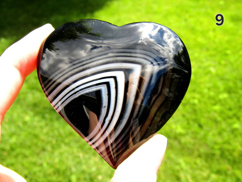 May include: A polished heart-shaped stone featuring a smooth, glossy black surface with distinct, concentric white and translucent gray banding patterns that form a sharp, angular chevron design across the center of the object.
