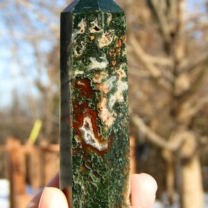 May include: A green and brown gemstone obelisk with a pointed top. The stone features a mix of green, brown, and white patterns. The obelisk is held in a hand against a blurred outdoor background.
