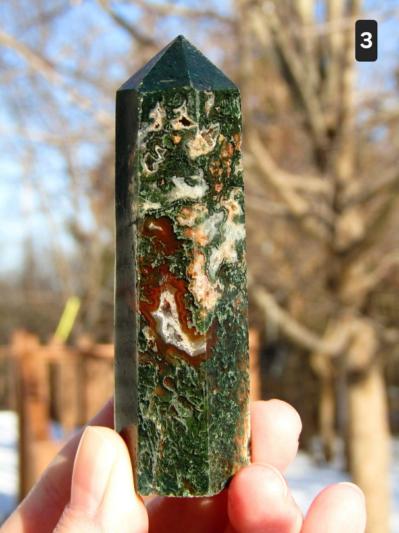 May include: A green and brown gemstone obelisk with a pointed top. The stone features a mix of green, brown, and white patterns. The obelisk is held in a hand against a blurred outdoor background.