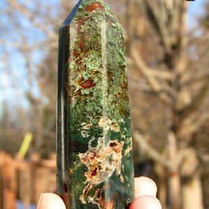 May include: A tall, polished gemstone obelisk. The stone features a mix of green, brown, and red hues, with a textured surface. The obelisk is held in a hand against a blurred outdoor background.