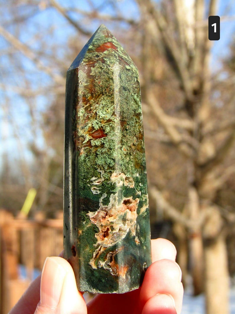 May include: A tall, polished gemstone obelisk. The stone features a mix of green, brown, and red hues, with a textured surface. The obelisk is held in a hand against a blurred outdoor background.