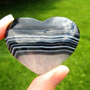 May include: A heart-shaped polished stone featuring horizontal bands of black, white, and translucent gray. The surface is smooth and reflective, displaying natural mineral layers. The number 3 is visible in the upper right corner of the image.