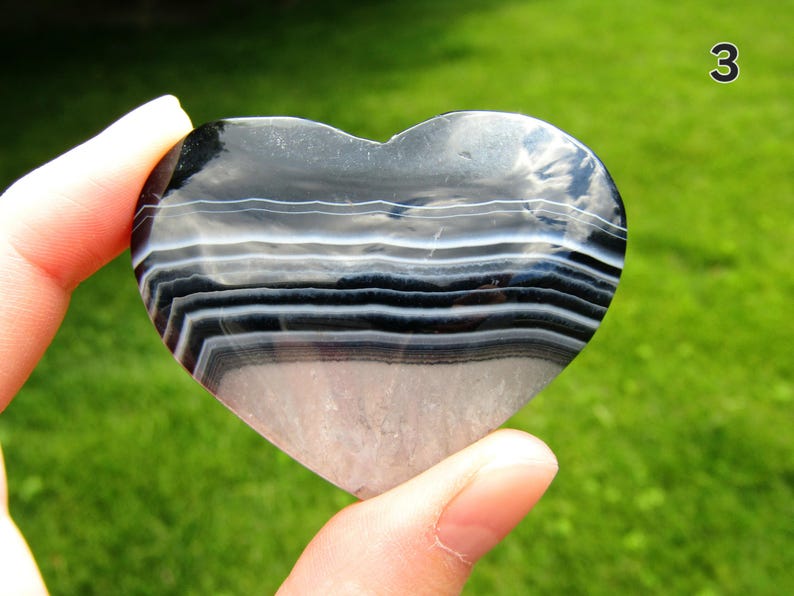 May include: A heart-shaped polished stone featuring horizontal bands of black, white, and translucent gray. The surface is smooth and reflective, displaying natural mineral layers. The number 3 is visible in the upper right corner of the image.