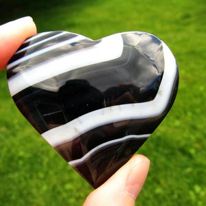 May include: A polished heart-shaped stone featuring alternating bands of deep black and opaque white. The smooth, glossy surface reflects light, highlighting the distinct, curved, and parallel striped patterns across the entire object.