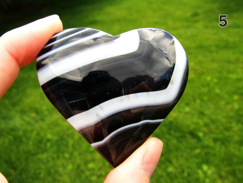 May include: A polished heart-shaped stone featuring alternating bands of deep black and opaque white. The smooth, glossy surface reflects light, highlighting the distinct, curved, and parallel striped patterns across the entire object.