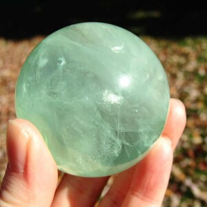 May include: A translucent, light green fluorite sphere is held in a hand. The crystal ball has internal veining and a smooth, polished surface. The background is blurred, with hints of brown and green.